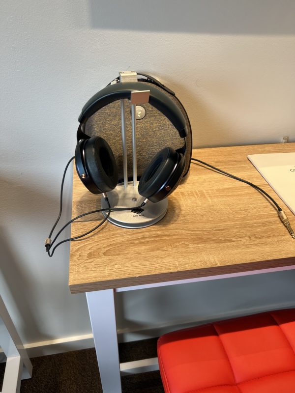 A pair of black over-ear headphones sits on a silver headphone stand placed on a wooden desk next to a closed laptop with a white cover.